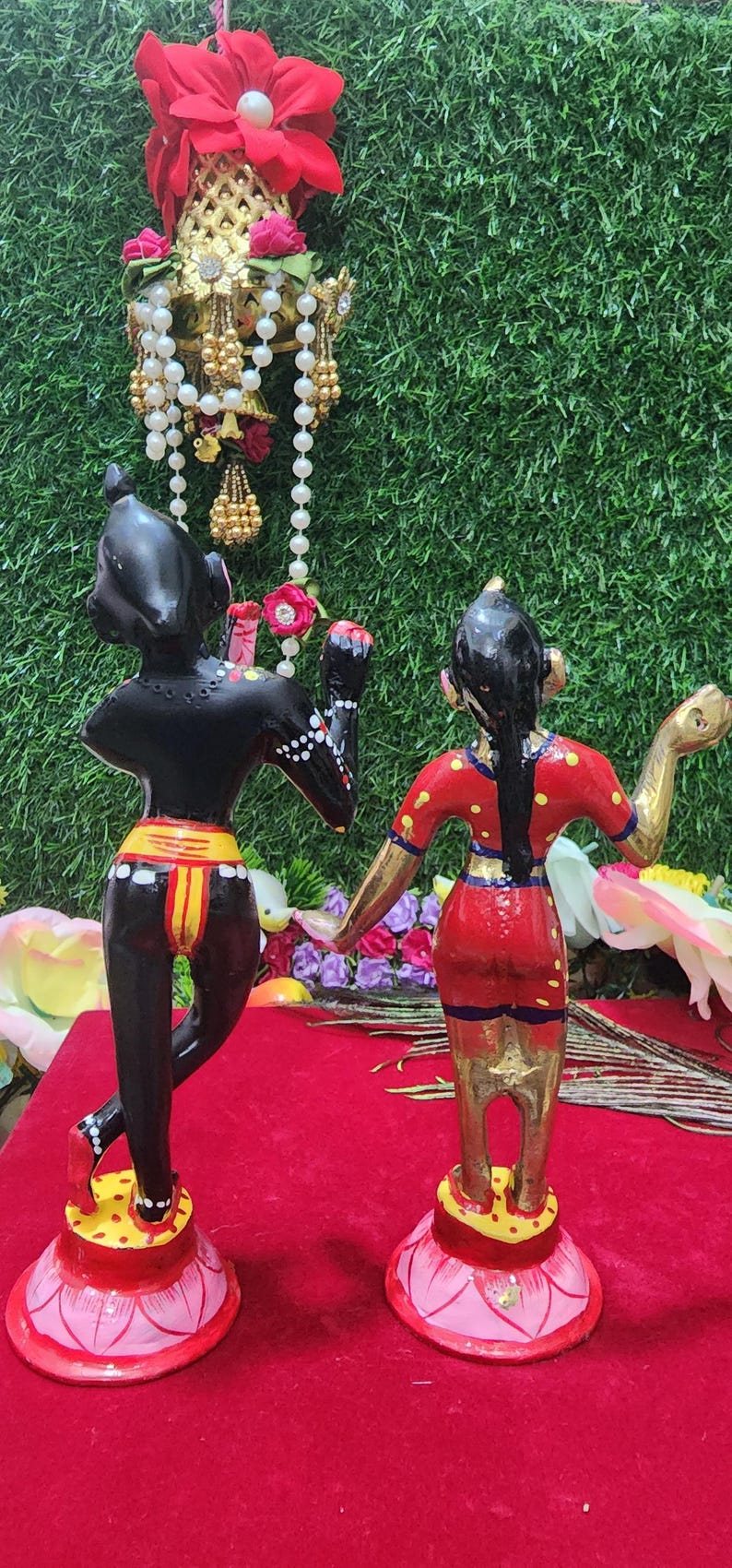 Op de afbeelding: Een paar handgeschilderde beeldjes die een stel in traditionele Indiase kleding voorstellen. Het mannelijke beeldje is zwart met gele en rode accenten, terwijl het vrouwelijke beeldje een rode jurk draagt met gele en blauwe accenten. Beide beeldjes staan op lotusvormige voetstukken.