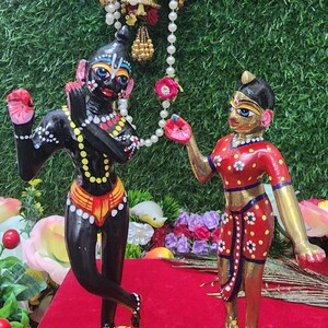Op de afbeelding: Handgeschilderde houten beeldjes van Radha en Krishna, staande op lotusvormige bases. Krishna is zwart met gele en witte accenten, terwijl Radha goudkleurig is met rode en witte accenten. Ze dragen allebei traditionele Indiase kleding.