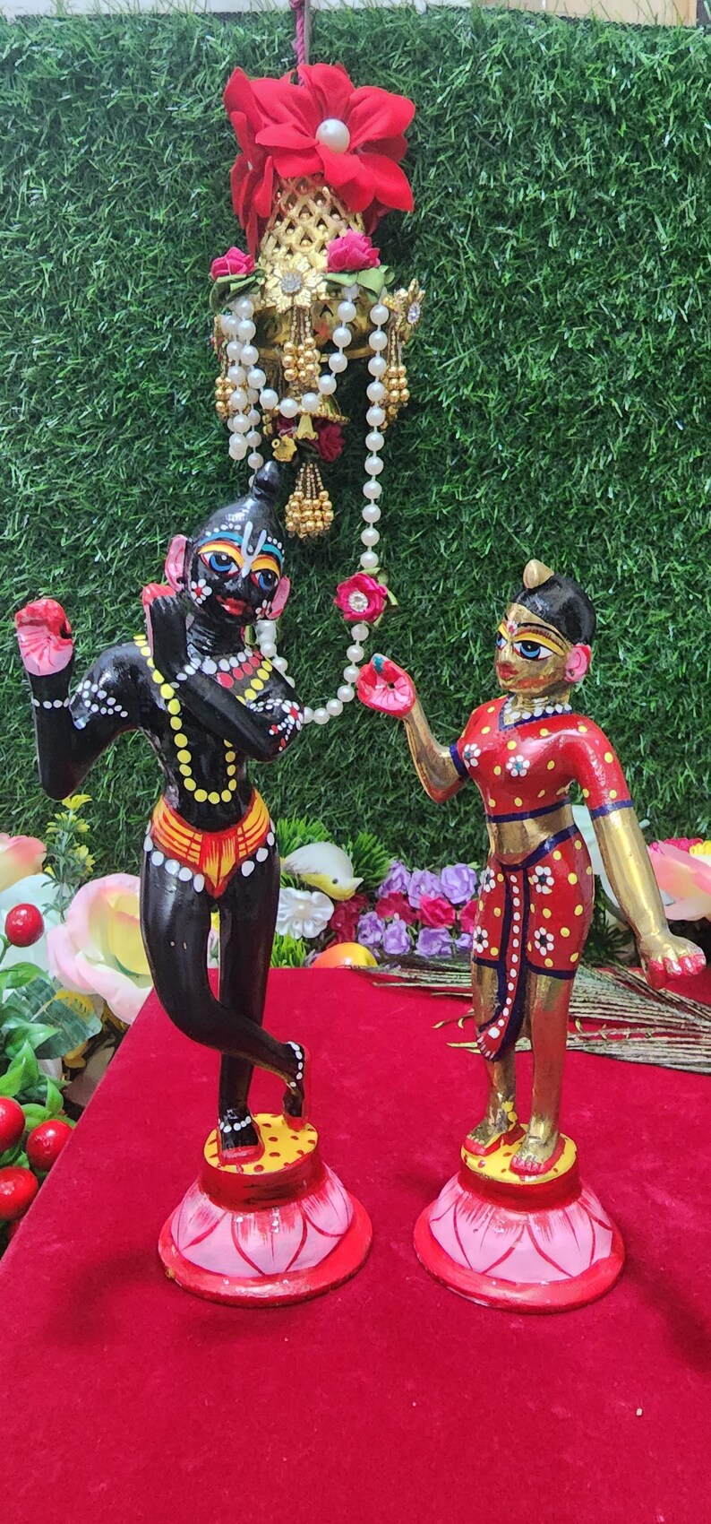Op de afbeelding: Handgeschilderde houten beeldjes van Radha en Krishna, staande op lotusvormige bases. Krishna is zwart met gele en witte accenten, terwijl Radha goudkleurig is met rode en witte accenten. Ze dragen allebei traditionele Indiase kleding.