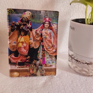 May include: A rectangular decorative panel featuring a colorful depiction of Hindu deities. The artwork showcases Krishna playing a flute, with Radha and other figures in traditional attire. The panel is set against a backdrop of a temple.