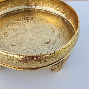 May include: A gold metal tray with an ornate design and a small elephant figure on the handle. The tray has three legs and is ideal for serving or displaying items.