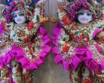 May include: Two ornate figurines of Hindu deities, dressed in colourful clothing with intricate details. The figures are standing on marble bases and are holding golden chains with bells.