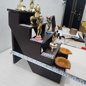 May include: A dark brown wooden tiered shelf with five levels, decorated with white and pink floral designs. The shelf is adorned with five gold-colored figurines and two brown clay diyas.