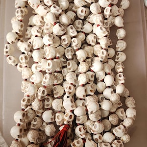 May include: A white skull bead necklace with red and black tassels. The beads are made of bone and have a cracked texture. The necklace is approximately 18 inches long.
