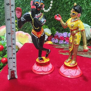 Op de afbeelding: Een paar handgeschilderde beeldjes die een stel in traditionele Indiase kleding voorstellen. Het mannelijke beeldje is zwart met gele en rode accenten, terwijl het vrouwelijke beeldje geel is met rode en gouden accenten. Beide beeldjes staan op lotusvormige bases.