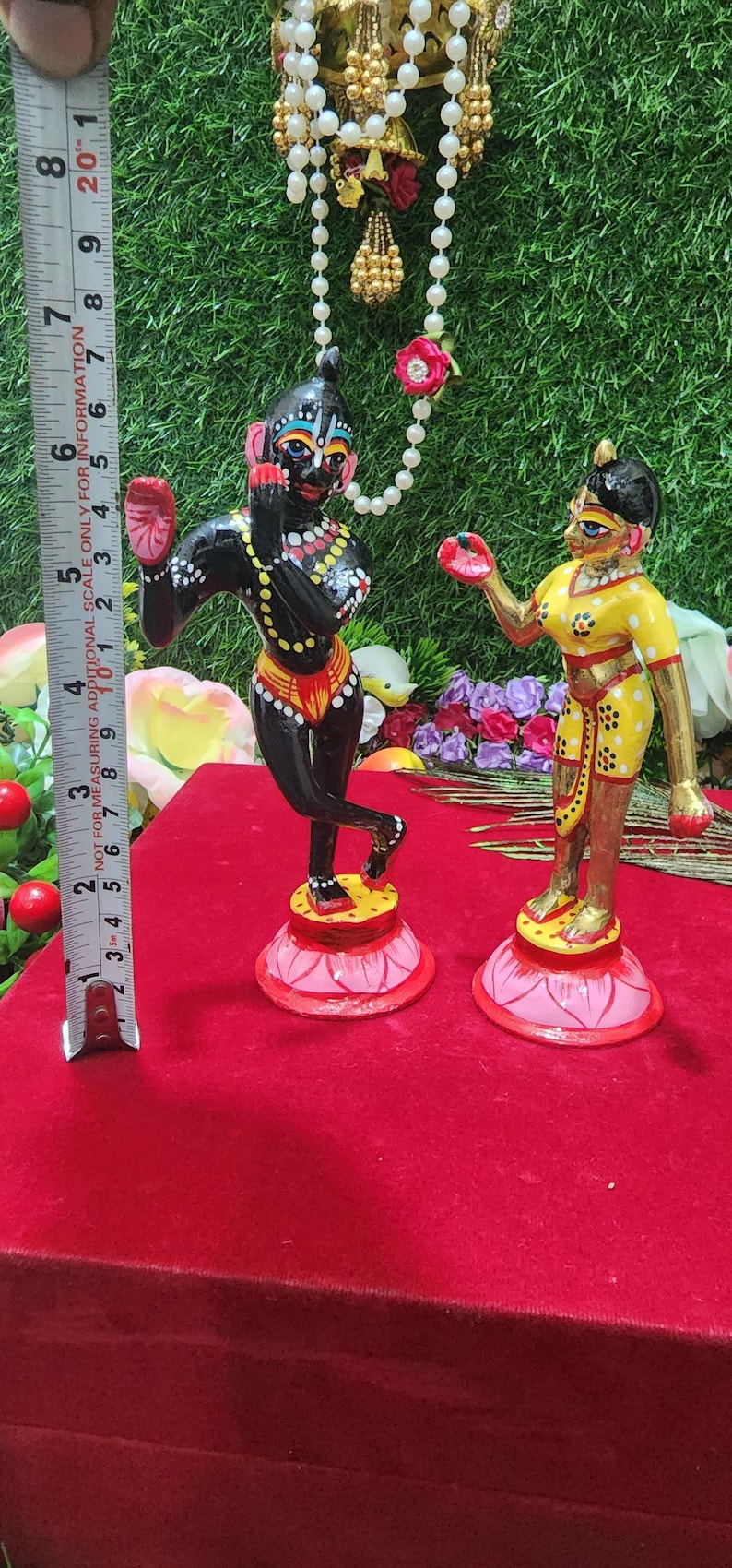Op de afbeelding: Een paar handgeschilderde beeldjes die een stel in traditionele Indiase kleding voorstellen. Het mannelijke beeldje is zwart met gele en rode accenten, terwijl het vrouwelijke beeldje geel is met rode en gouden accenten. Beide beeldjes staan op lotusvormige bases.