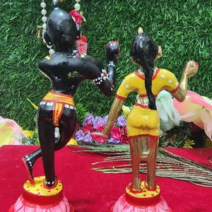 Op de afbeelding: Een paar handgeschilderde houten beeldjes die een stel in een traditionele Indiase danspose voorstellen. De mannelijke figuur is zwart met witte stippen en de vrouwelijke figuur is geel met witte stippen. Beide figuren staan op lotusvormige bases.