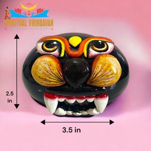 May include: A black wooden mask with a fierce expression. The mask has yellow and orange painted details, including eyes, nose, and teeth. The mask is 3.5 inches wide and 2.5 inches tall.