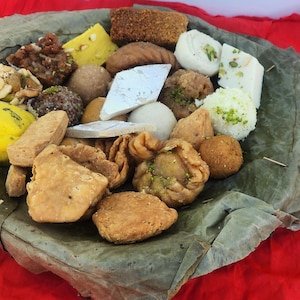 May include: A variety of Indian sweets and snacks arranged on a large green leaf. The sweets include ladoo, barfi, and jalebi. The snacks include samosas and pakoras.
