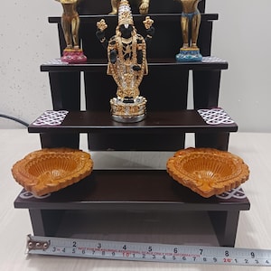 May include: A dark brown wooden tiered shelf with three levels, each decorated with a white and black geometric pattern. The top level features a gold-colored statue of a deity with a black face and a golden crown. The middle level has two gold-colored statues of deities with raised arms. The bottom level has two brown clay diya lamps.
