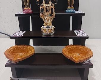 May include: A dark brown wooden tiered shelf with three levels, each decorated with a white and black geometric pattern. The top level features a gold-colored statue of a deity with a black face and a golden crown. The middle level has two gold-colored statues of deities with raised arms. The bottom level has two brown clay diya lamps.
