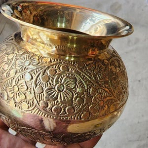 May include: A gold metal pot with intricate floral designs etched into the surface. The pot has a wide mouth and a rounded body.