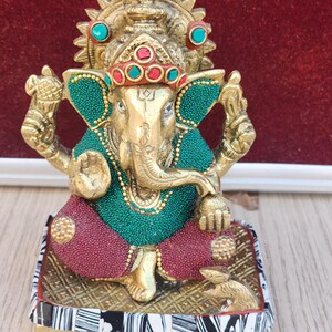 Może przedstawiać: Złota statuetka Ganesha, hinduskiego boga mądrości i dobrobytu, z zielonymi i czerwonymi akcentami. Statuetka stoi na kwadratowej podstawie z czarno-białym wzorem.