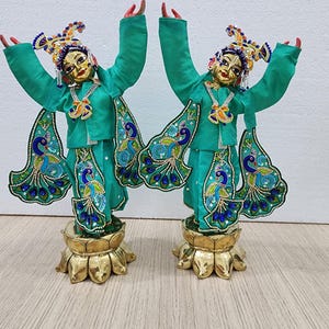 May include: Two teal blue and gold statues of figures with peacock feather designs and elaborate headwear. The figures are standing on gold lotus-shaped bases.