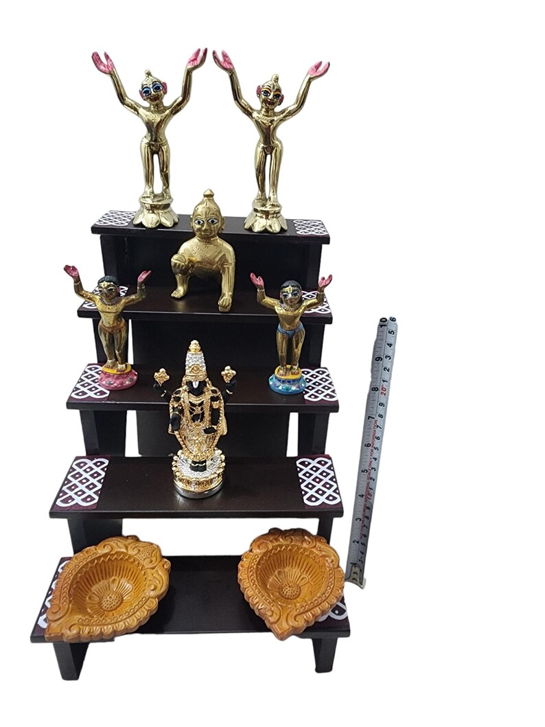 May include: A three-tiered wooden shelf with decorative details, featuring five gold and colorful figurines, including two standing figures with raised arms, a seated figure, and two standing figures with one hand raised. The shelf also includes two decorative diyas, or oil lamps, on the bottom tier.