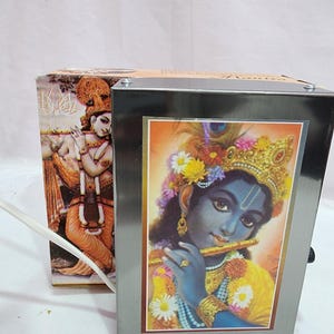 May include: A silver metal frame with a picture of Krishna playing a flute. The picture is surrounded by a gold border and has a floral design. The frame is on a white surface.