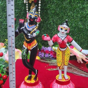 Op de afbeelding: Twee handgeschilderde beeldjes van hindoe&iuml;stische goden, Krishna en Radha, staand op lotusvormige bases. Krishna speelt fluit en Radha houdt een bloem vast. De beeldjes zijn geschilderd in levendige kleuren, waaronder zwart, rood, geel en groen.