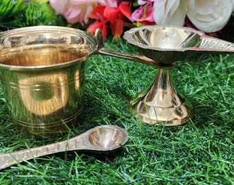 May include: A set of three brass puja items, including a small cup, a diya, and a spoon. The items are arranged on a green artificial grass surface.