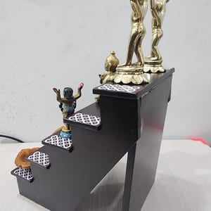 May include: A wooden tiered stand with four steps, each decorated with a white and black geometric pattern. The top step features two gold figurines with raised arms, and a smaller gold figurine on the step below.