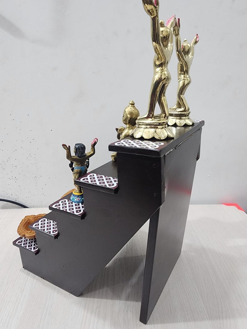 May include: A wooden tiered stand with four steps, each decorated with a white and black geometric pattern. The top step features two gold figurines with raised arms, and a smaller gold figurine on the step below.