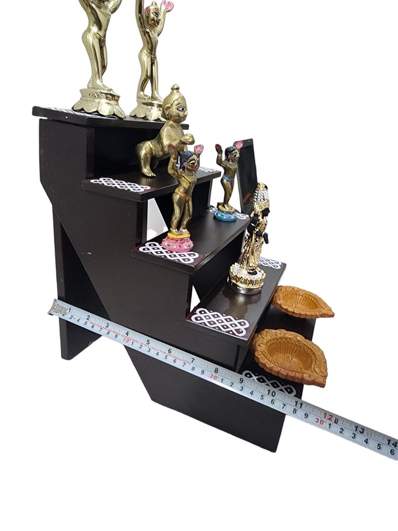 May include: A four-tiered wooden shelf with decorative details, featuring a variety of small gold and colorful figurines, including a baby Krishna, and two diya oil lamps.