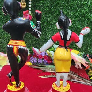Op de afbeelding: Een paar handgeschilderde beeldjes die een stel in traditionele Indiase kleding voorstellen. De mannelijke figuur is zwart met gele en rode accenten, terwijl de vrouwelijke figuur wit is met rode, gele en zwarte accenten. Beide figuren staan op roze lotusbases.