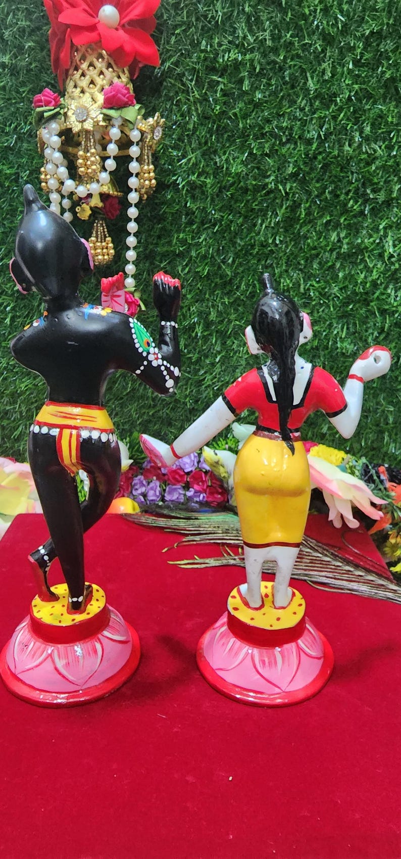 Op de afbeelding: Een paar handgeschilderde beeldjes die een stel in traditionele Indiase kleding voorstellen. De mannelijke figuur is zwart met gele en rode accenten, terwijl de vrouwelijke figuur wit is met rode, gele en zwarte accenten. Beide figuren staan op roze lotusbases.