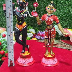 Op de afbeelding: Een paar handgeschilderde beeldjes die een stel voorstellen dat op lotusvormige bases staat. De mannelijke figuur is zwart met gele en rode accenten, terwijl de vrouwelijke figuur goudkleurig is met rode en witte accenten. Beide figuren houden een touwtje met witte kralen vast.