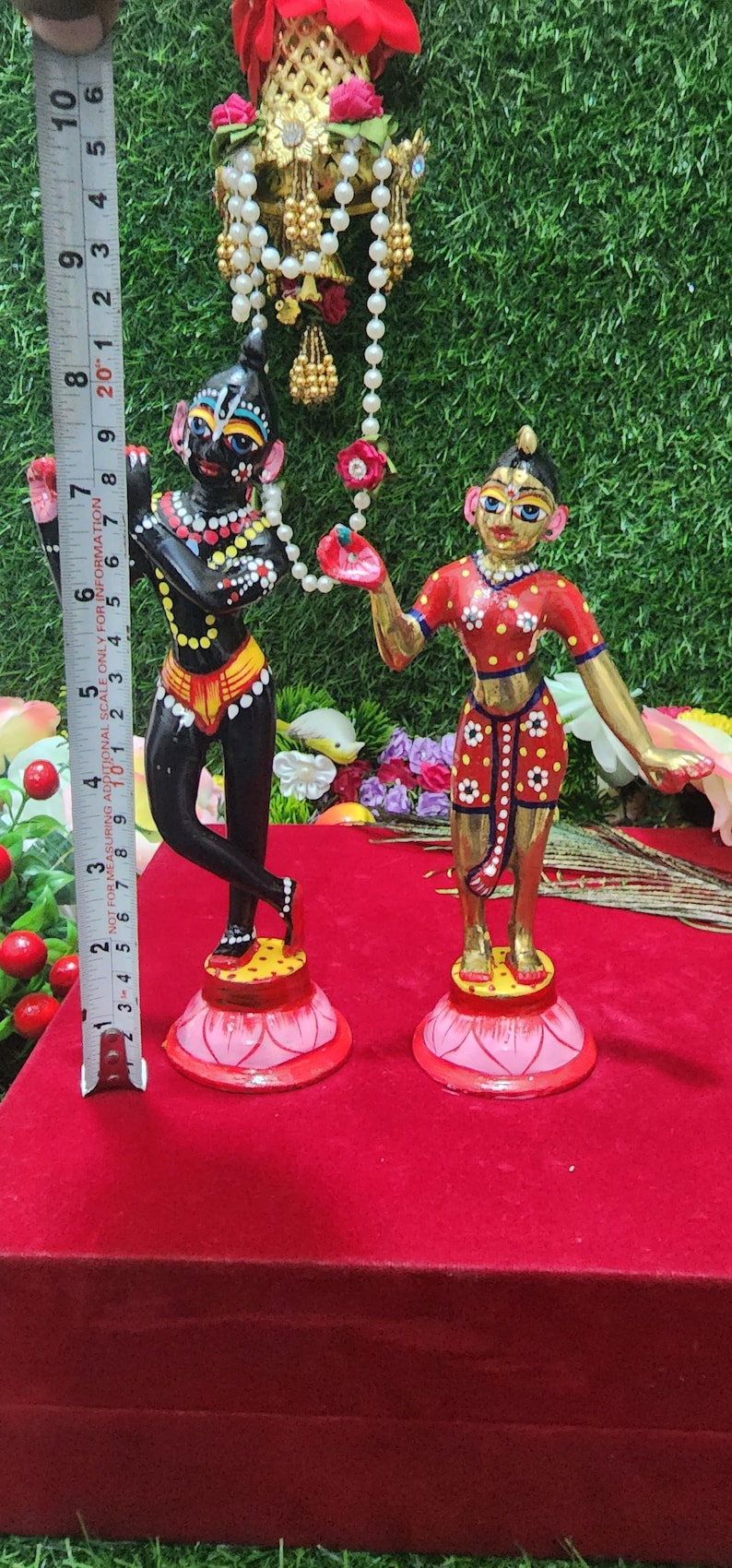 Op de afbeelding: Een paar handgeschilderde beeldjes die een stel voorstellen dat op lotusvormige bases staat. De mannelijke figuur is zwart met gele en rode accenten, terwijl de vrouwelijke figuur goudkleurig is met rode en witte accenten. Beide figuren houden een touwtje met witte kralen vast.