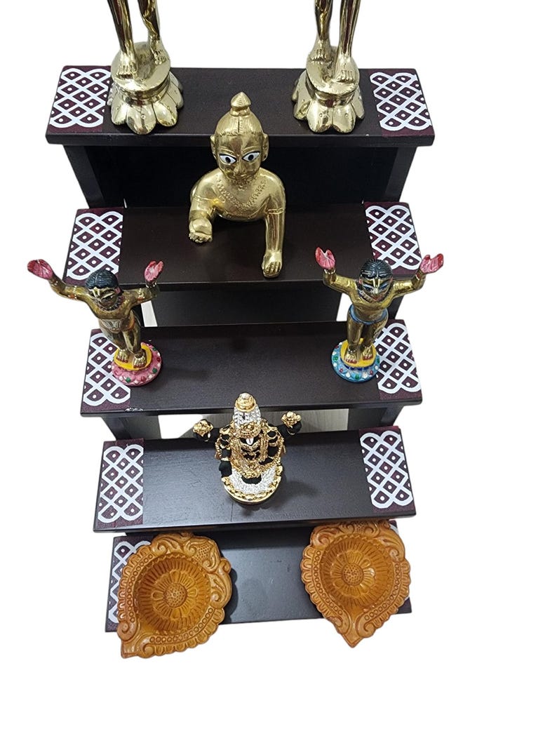 May include: A four-tiered wooden shelf with decorative details, featuring gold and silver figurines of deities, including a baby Krishna, and two diya lamps.