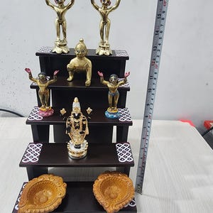 May include: A three-tiered wooden shelf with decorative details, featuring five gold and blue figurines, and two brown diya lamps. The shelf is approximately 17 inches tall.