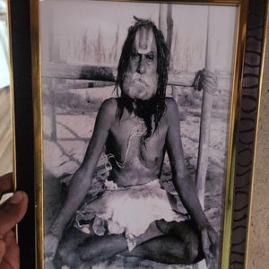 Puede incluir: Una fotografía en blanco y negro de un hombre sentado en una postura meditativa. Tiene el pelo largo y barba, y lleva una tela blanca alrededor de la cintura. La foto está enmarcada en un marco negro y dorado.