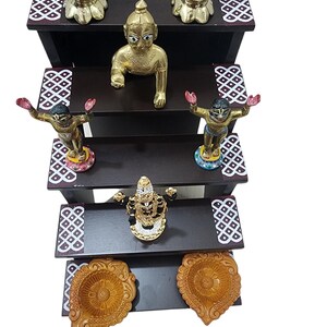 May include: A four-tiered wooden shelf with decorative details, featuring gold and silver figurines of deities, including a baby Krishna, and two diya lamps.
