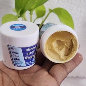 Puede incluir: Un tarro de plástico blanco con una etiqueta azul que dice "Shri Dham Gopi Tilak A Quality Product INDIA". El tarro contiene una pasta amarilla. El tarro también está etiquetado en hindi.