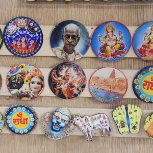 Op de afbeelding: Een verzameling ronde magneten en decoratieve items met religieuze figuren, goden en culturele symbolen. De magneten zijn gerangschikt op houten strips en bevatten verschillende kleuren en ontwerpen.
