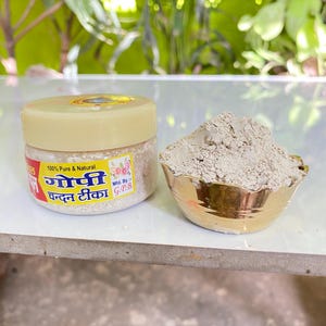 Może przedstawiać: Żółty plastikowy pojemnik z białą pokrywką i etykietą z napisem "100% Pure & Natural गोपी चक्कन टिका Mfd. By: G.P.B.". Pojemnik jest wypełniony białym proszkiem. Obok pojemnika znajduje się mała złota miska wypełniona tym samym białym proszkiem.