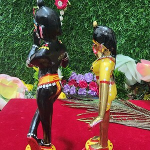 Op de afbeelding: Een paar handgeschilderde houten beeldjes die een stel voorstellen dat op lotusvormige bases staat. De mannelijke figuur is zwart met gele en witte accenten, terwijl de vrouwelijke figuur geel is met witte en gouden accenten. De bases zijn rood geverfd met roze lotusbloemen.