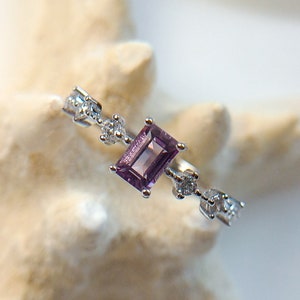Peut inclure: Une bague en argent ornée d'une pierre précieuse rectangulaire violette. La bague est ornée de plus petites pierres précieuses claires le long de la bande. La bague est présentée sur une étoile de mer blanche.