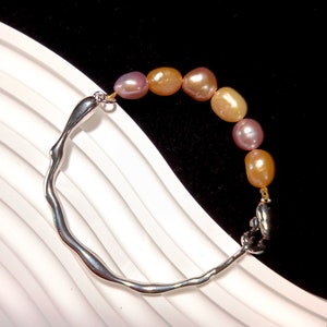 May include: A silver bracelet with a textured, irregular shape and a clasp. The bracelet is adorned with a strand of pastel pink and peach pearls.