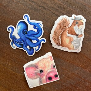 Op de afbeelding: Drie aquarel stickers met een blauwe octopus, een bruine eekhoorn met een hoed en een roze varken. De stickers zijn op een bruin houten oppervlak.
