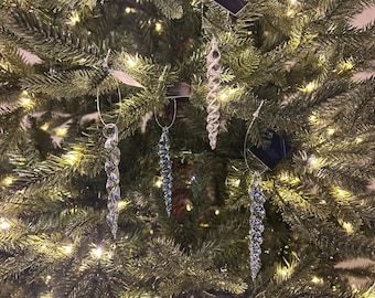 May include: A selection of clear, decorative icicle ornaments suspended from a Christmas tree. The ornaments feature spiral or faceted designs and are hung by silver wire. The tree is illuminated with small, warm white lights.