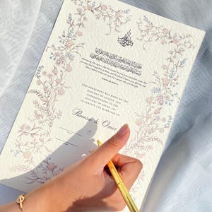 May include: A cream-coloured marriage certificate with floral border and Arabic calligraphy. The document includes text in English and Arabic, with the names Raeesa & Omer. A gold pen is held over the document.