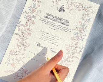 Könnte beinhalten: Eine cremefarbene Heiratsurkunde mit floralem Rand und arabischer Kalligraphie. Das Dokument enthält Text in Englisch und Arabisch mit den Namen Raeesa & Omer. Ein goldener Stift wird über dem Dokument gehalten.