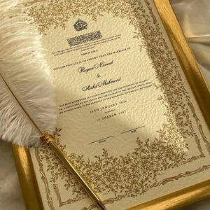 Peut inclure: Certificat de mariage encadré avec bordure florale dorée et texte. Le certificat est pour Rayan Naveed et Aisha Mahmood, daté du 29 janvier 2026. Un stylo doré et une plume blanche sont posés sur le certificat.