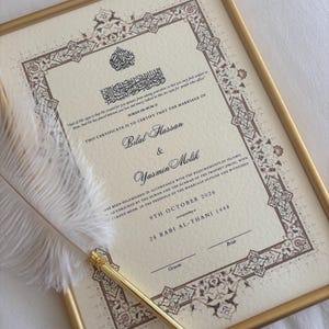 May include: A framed Islamic marriage certificate with cream-colored paper, ornate brown and gold borders, and black calligraphy. The text includes the names Bilal Hassan and Yasmin Molik, dated 9th October 2026. A white feather quill pen rests on top.