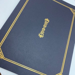 Puede incluir: Una carpeta azul oscuro con un borde dorado y un diseño decorativo dorado en el centro.