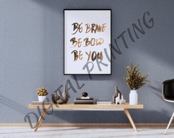 May include: A framed print with the words "BE BRAVE BE BOLD BE YOU" in gold script hangs above a wooden table. The table holds decorative items, including a small potted plant and a vase with dried flowers. The wall is painted a muted blue.