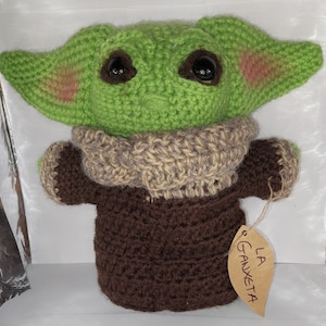 Puede incluir: Un muñeco de bebé Yoda de crochet verde que lleva un suéter de crochet marrón y beige. El muñeco tiene ojos grandes y una boca pequeña. El muñeco sostiene una etiqueta marrón con el texto "LA GAUXETA".