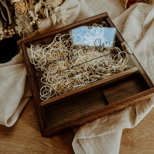 Könnte beinhalten: Eine dunkle Holz-Andenkenbox mit Glasdeckel, gefüllt mit dekorativem Garn. Auf der Box stehen die Namen "Jennifer & Igor" und das Wort "WEDDING" in eleganter Schrift. Ein kleines Holzfach ist im Inneren sichtbar.