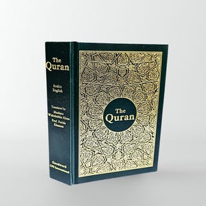 Op de afbeelding: Een groen hardback boek met gouden letters met de titel "De Koran" met Arabische en Engelse tekst. De omslag heeft een ingewikkeld gouden ontwerp.
